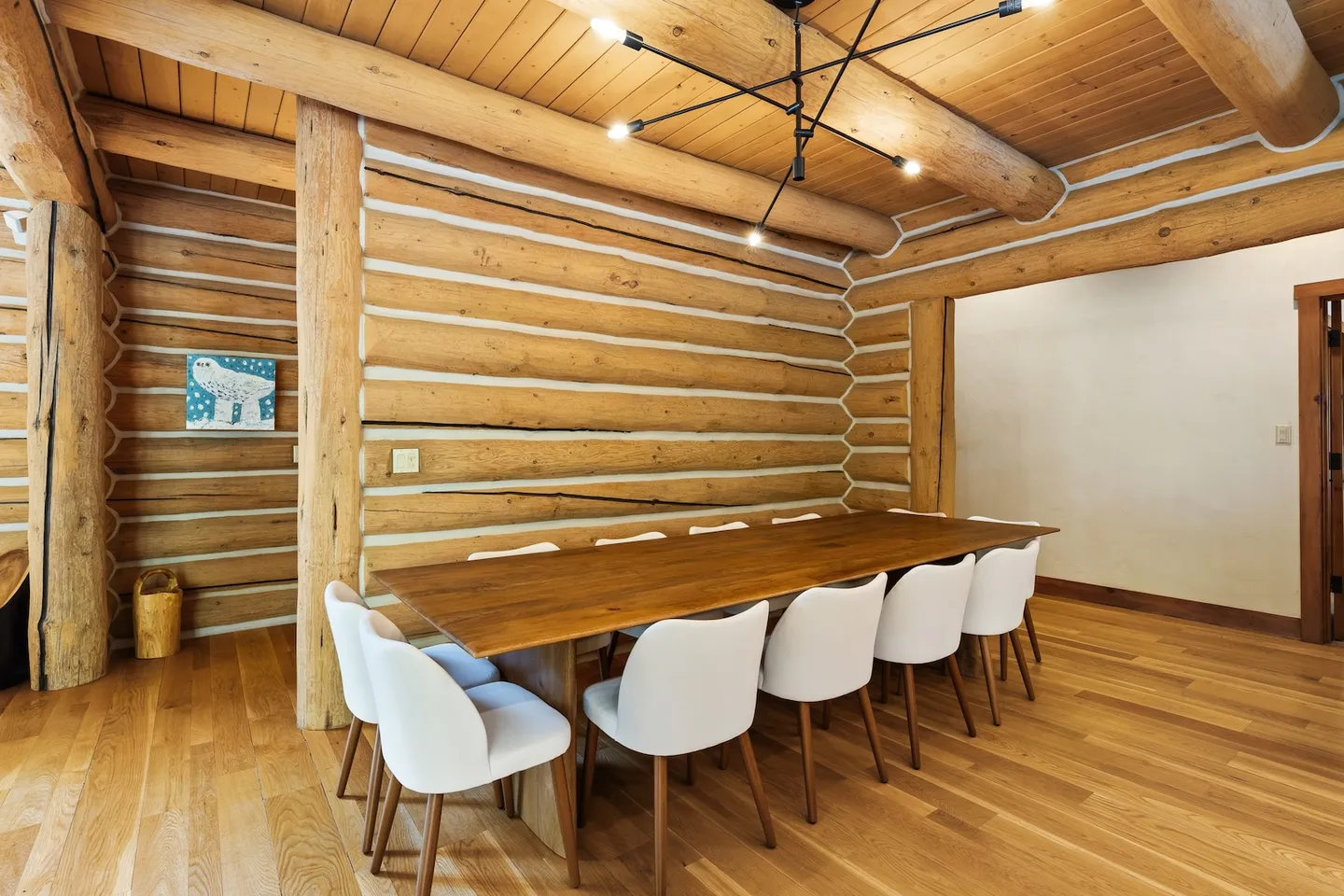Dining area with a large wooden table and white chairs in a room with exposed log walls and ceiling beams.
