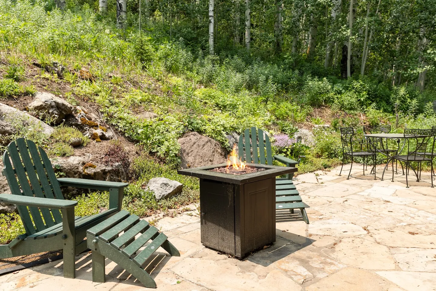 Outdoor stone patio with green Adirondack chairs around a lit gas fire pit backed by a wooded hillside.