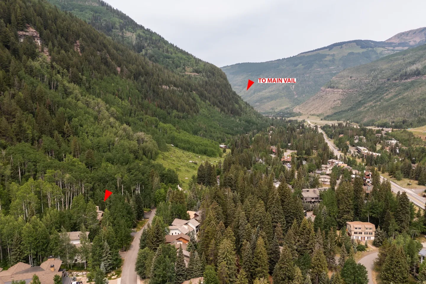 Aerial view of a mountain valley community surrounded by evergreen and deciduous trees, with steep mountainsides under an overcast sky.