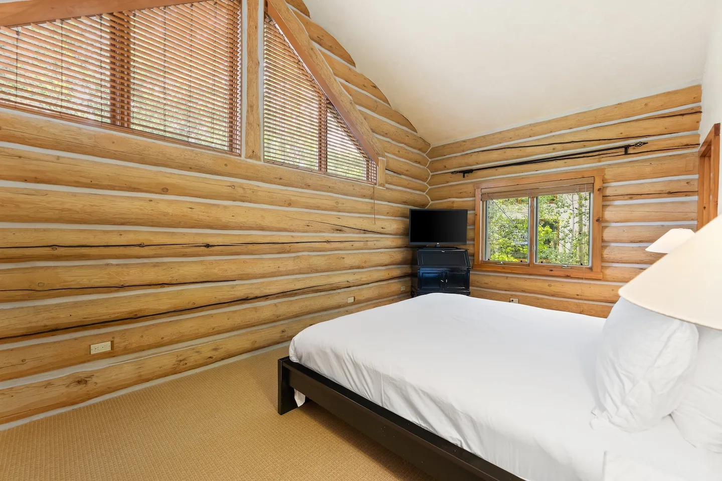 Bedroom with log cabin walls, vaulted ceiling with upper windows, and a bed made with white linens.
