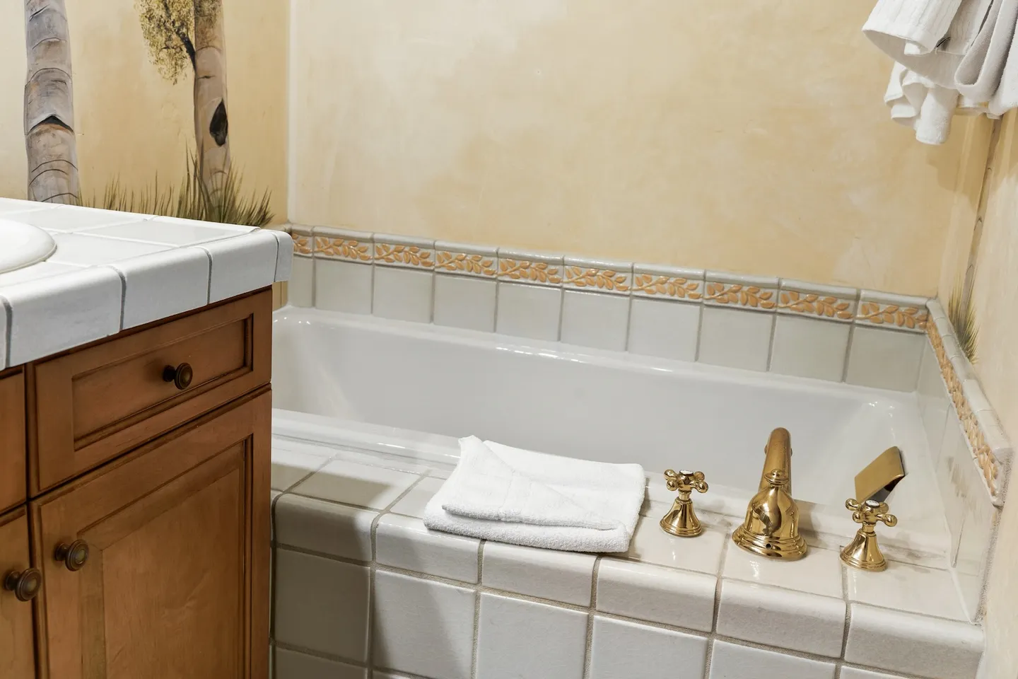 A tiled bathtub with brass fixtures next to a wooden vanity cabinet in a bathroom with painted birch tree wall art.