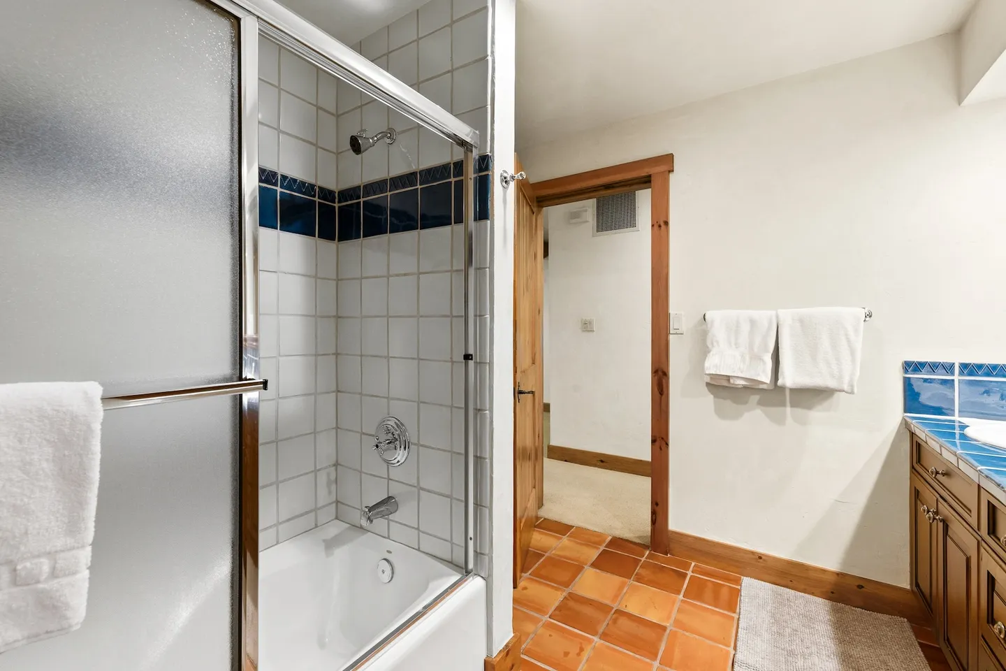 Bathroom with terra-cotta tile floor, shower-tub combo behind sliding glass doors, white walls, and wood vanity.
