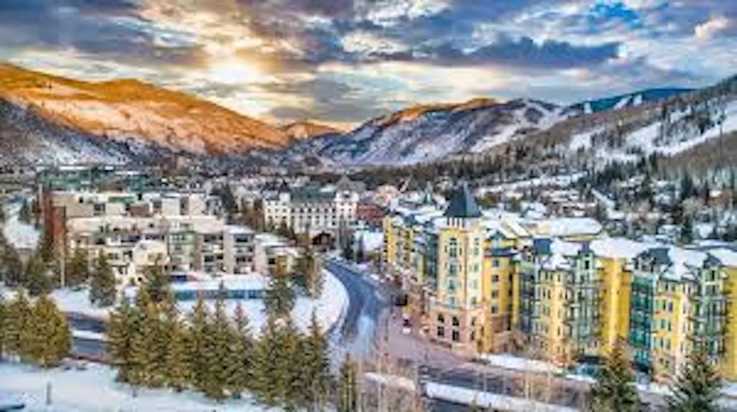 Elevated view of a mountain resort village with multistory lodge buildings and snow-covered slopes under a sunrise or sunset sky.