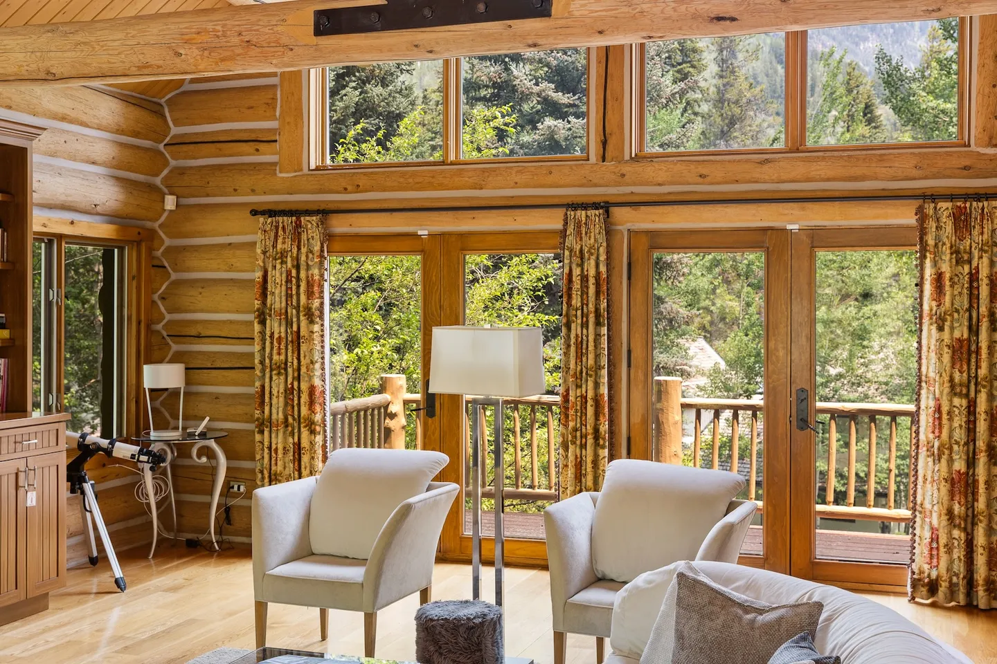 Living room in a log cabin featuring two light-colored armchairs, a floor lamp, and large windows looking out to an outdoor balcony and trees.