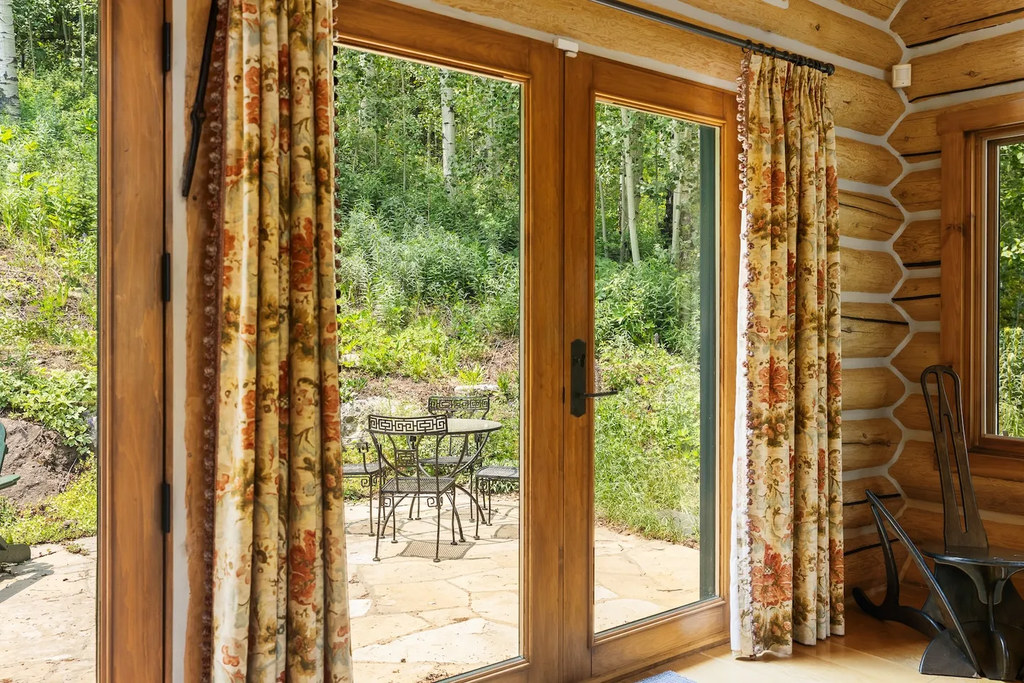 Glass double doors framed by wood trim and patterned curtains looking out to a stone patio with outdoor seating surrounded by trees.