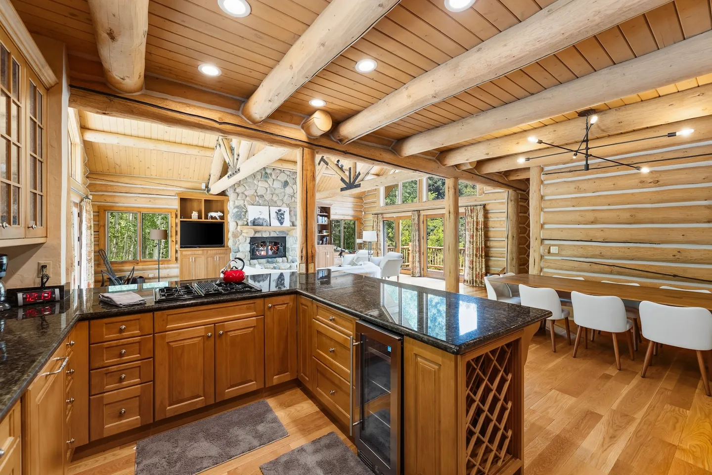 Open-concept log cabin interior showing a kitchen with dark countertops in the foreground, adjacent dining table, and living room with stone fireplace in the background.