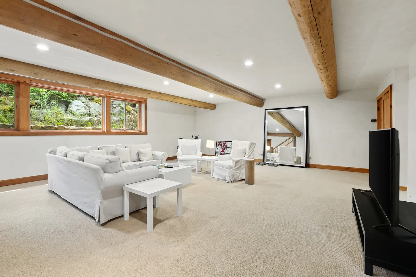 Spacious carpeted living room with white slipcovered seating, exposed ceiling log beams, and a television.