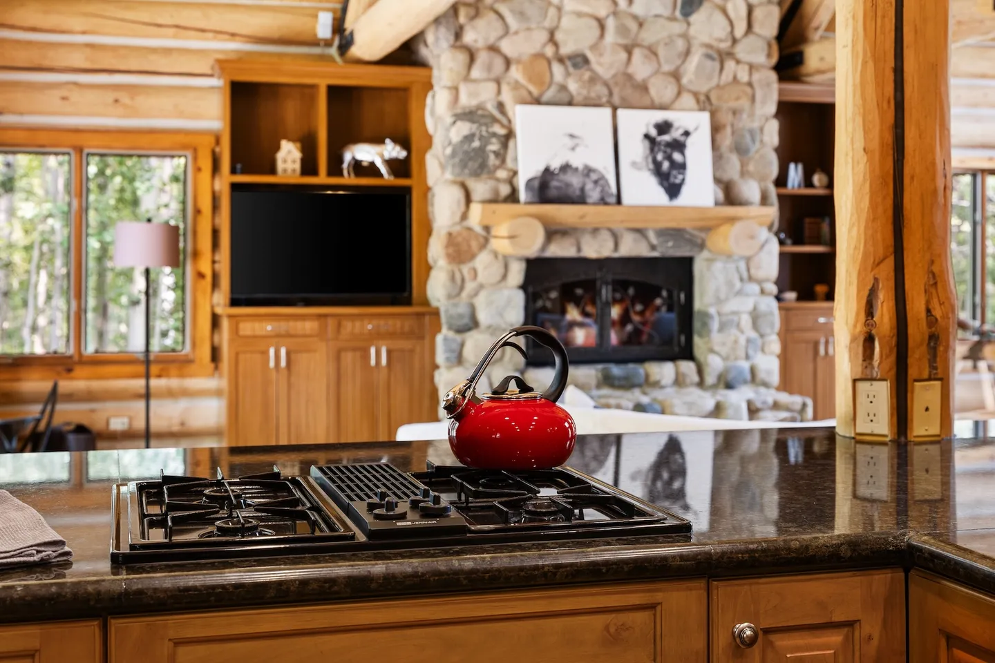 A red tea kettle on a dark countertop gas range, looking past log cabin walls toward a stone fireplace and television.