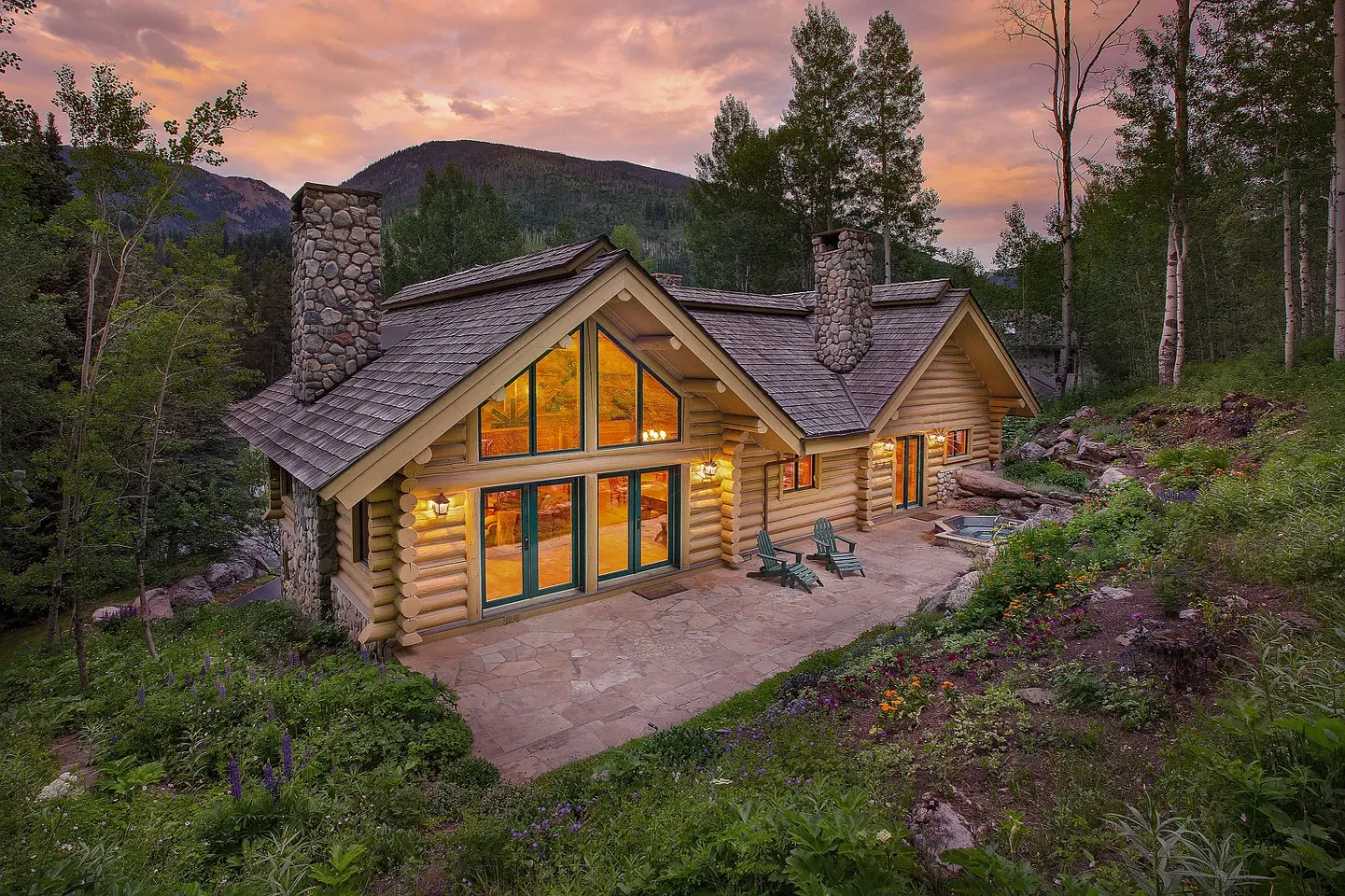 Exterior of a log home nestled on a hillside with stone chimneys, large windows, a stone patio, and an outdoor hot tub at dusk.