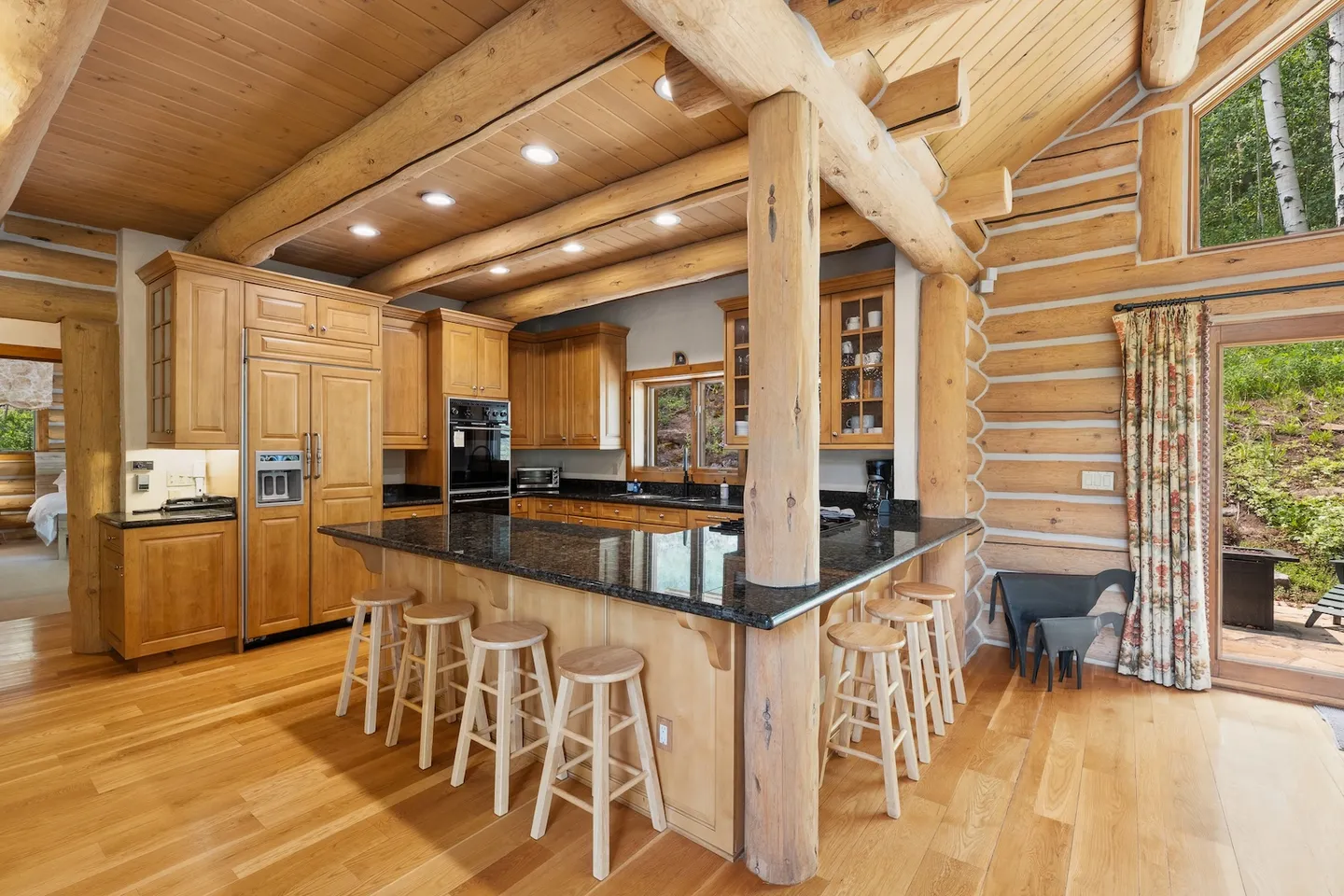 Log cabin kitchen with light wood cabinetry, dark granite countertops, and breakfast bar with wooden stools beneath a beamed log ceiling.