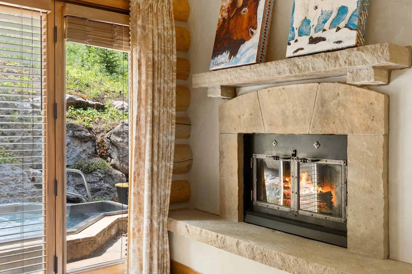 Stone fireplace with a lit fire next to a glass door looking out onto an outdoor hot tub and rocky hillside.