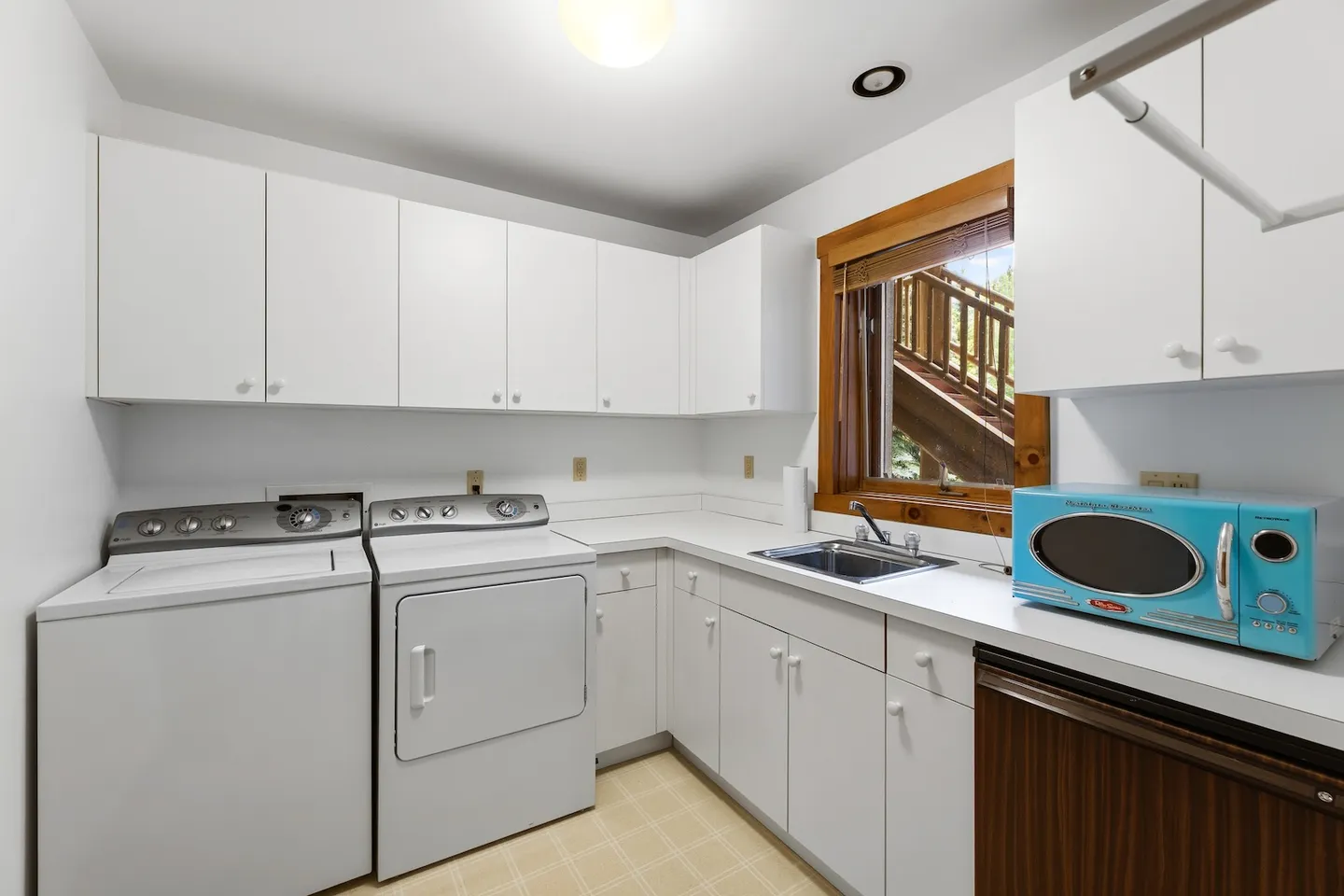 A laundry room with a white washer and dryer, white cabinets, a sink, and a blue microwave on the counter.