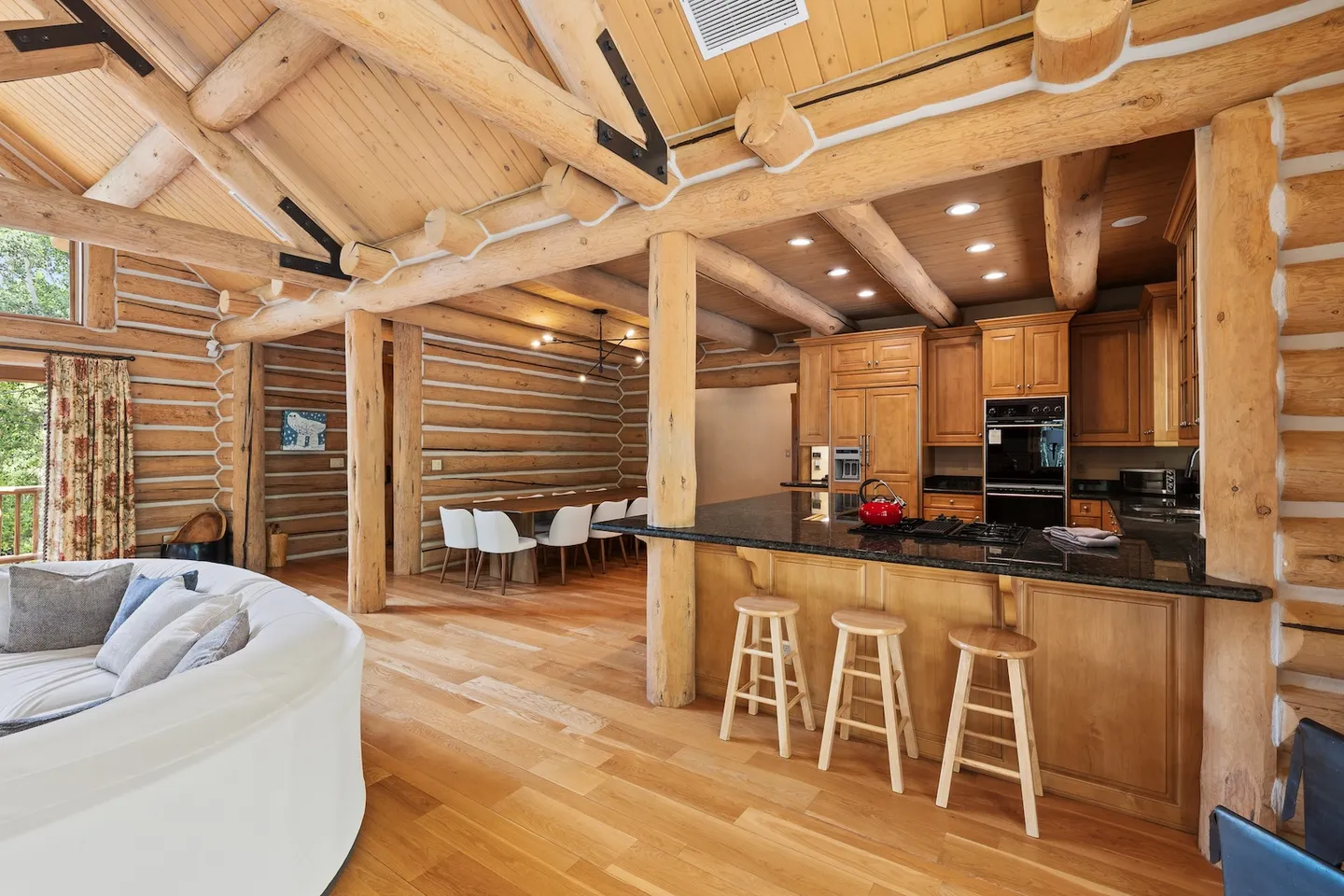 Open-concept log cabin interior featuring a kitchen with breakfast bar, dining area, and living space with wood log beams.