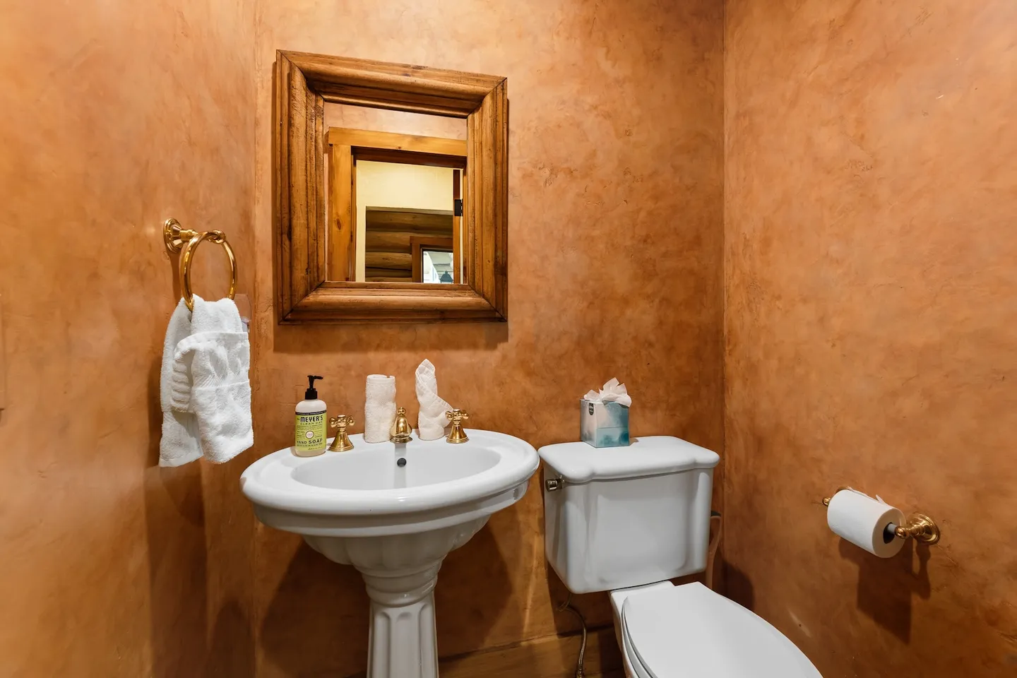 Powder room featuring textured warm terracotta walls, a white pedestal sink with brass fixtures, a toilet, and a wood-framed mirror.
