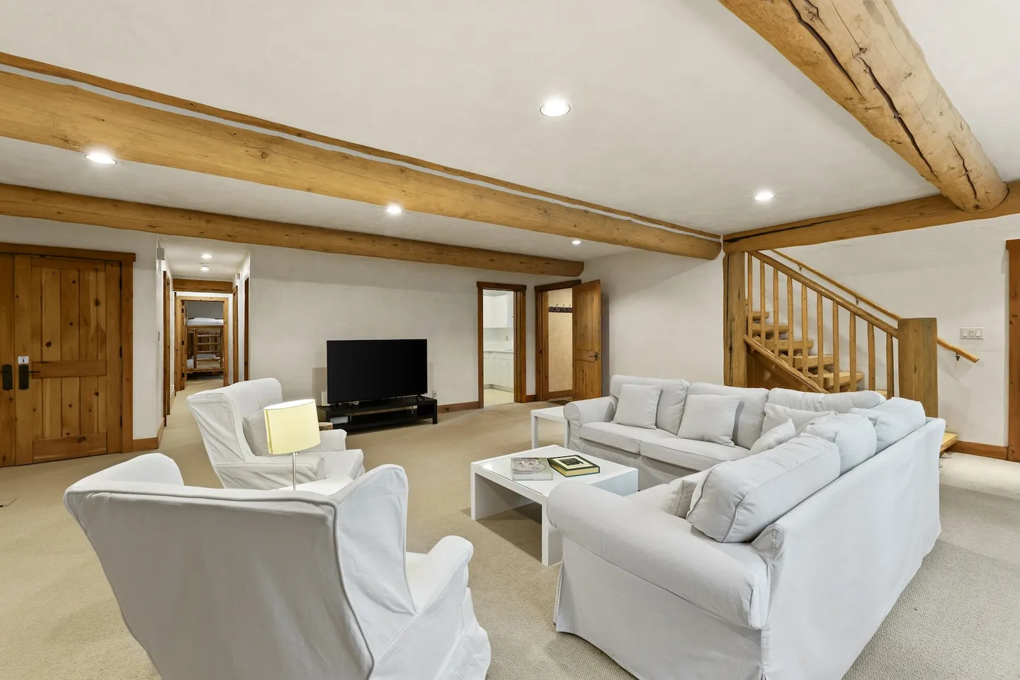 Spacious living area with exposed wood ceiling beams, white slipcovered seating, a TV, and stairs leading up.