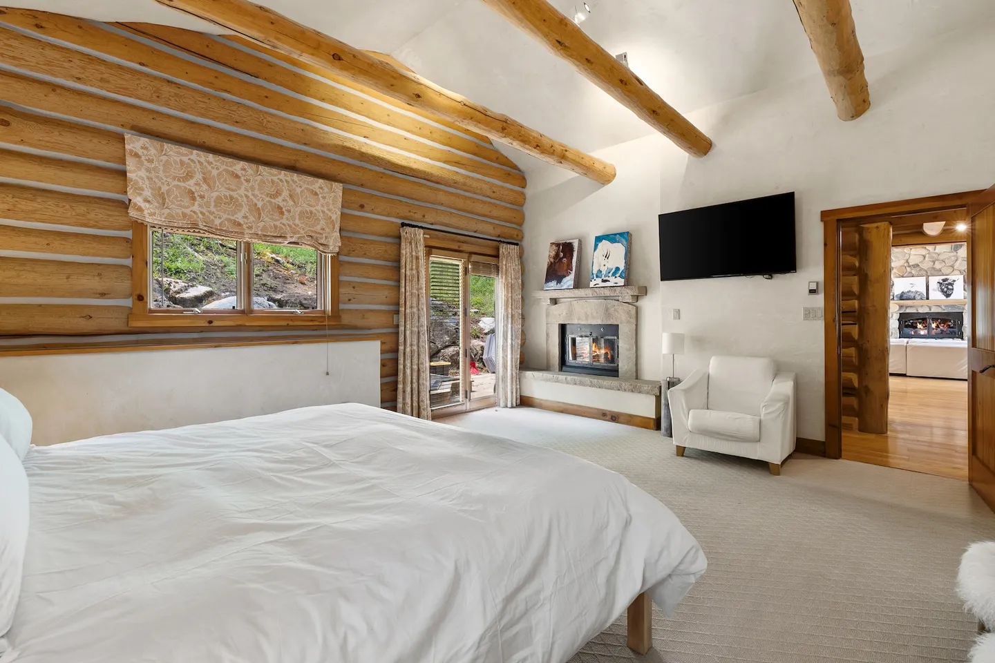 Bedroom with log walls, exposed log ceiling beams, a bed made with white linens, a fireplace, and a mounted TV.
