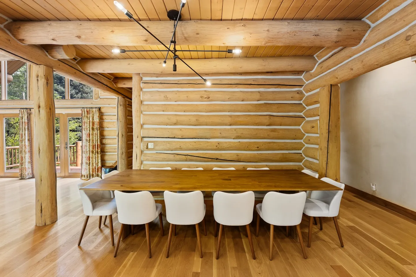 Dining area with a large wooden table, white upholstered chairs, log walls, and wood-beam ceiling.