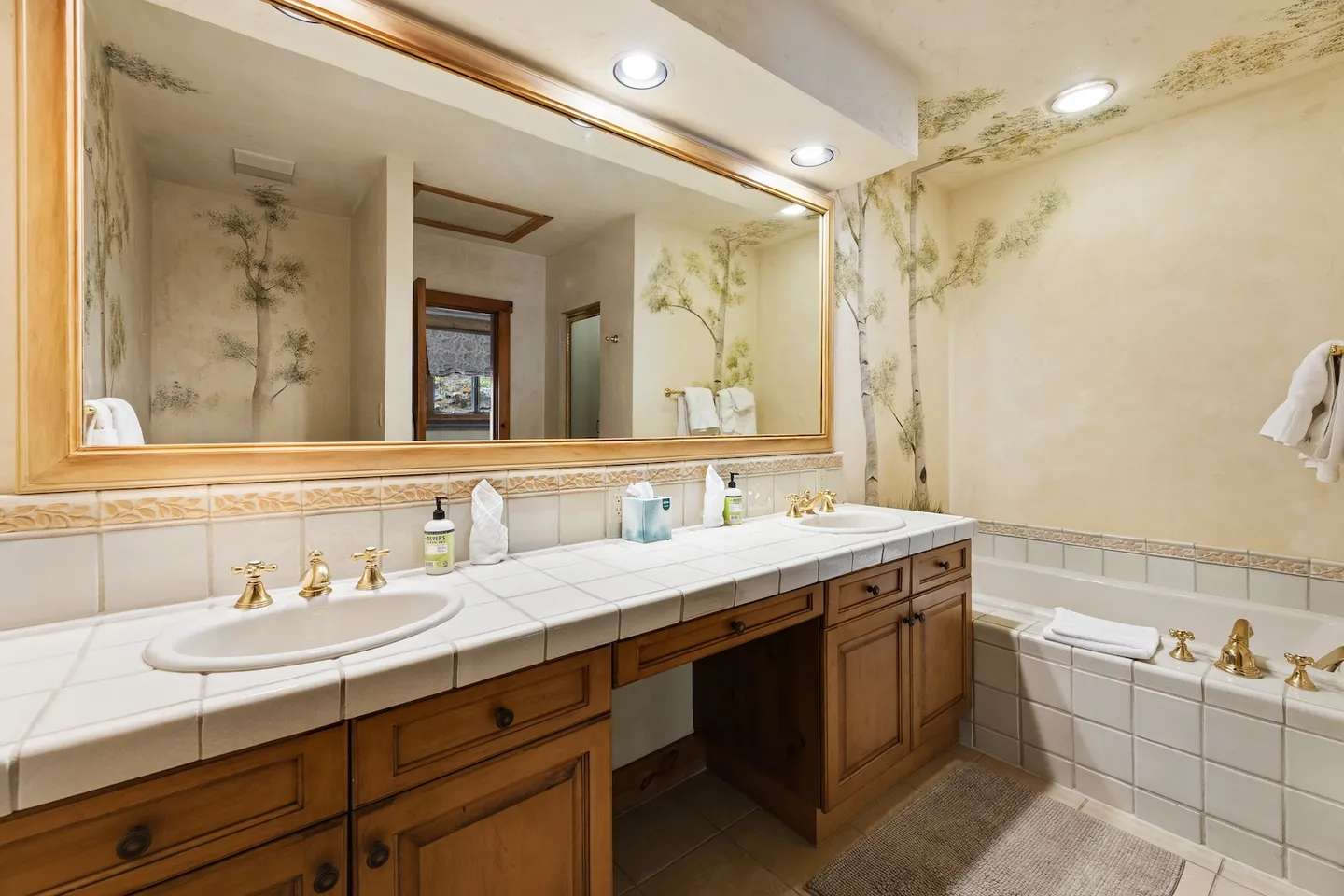 Bathroom with a double-sink tiled vanity, a large framed mirror, painted tree murals on the walls, and a built-in tiled bathtub.