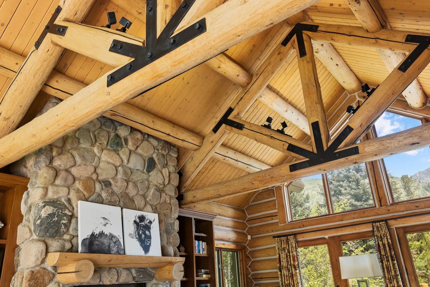 Upward view of a vaulted log ceiling with heavy wooden trusses, black metal brackets, and a stone fireplace.