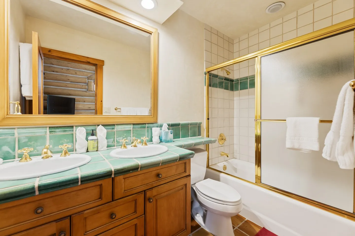 Bathroom with double vanity featuring green tile countertops, gold-trimmed mirror, toilet, and combination shower and bathtub with sliding glass doors.