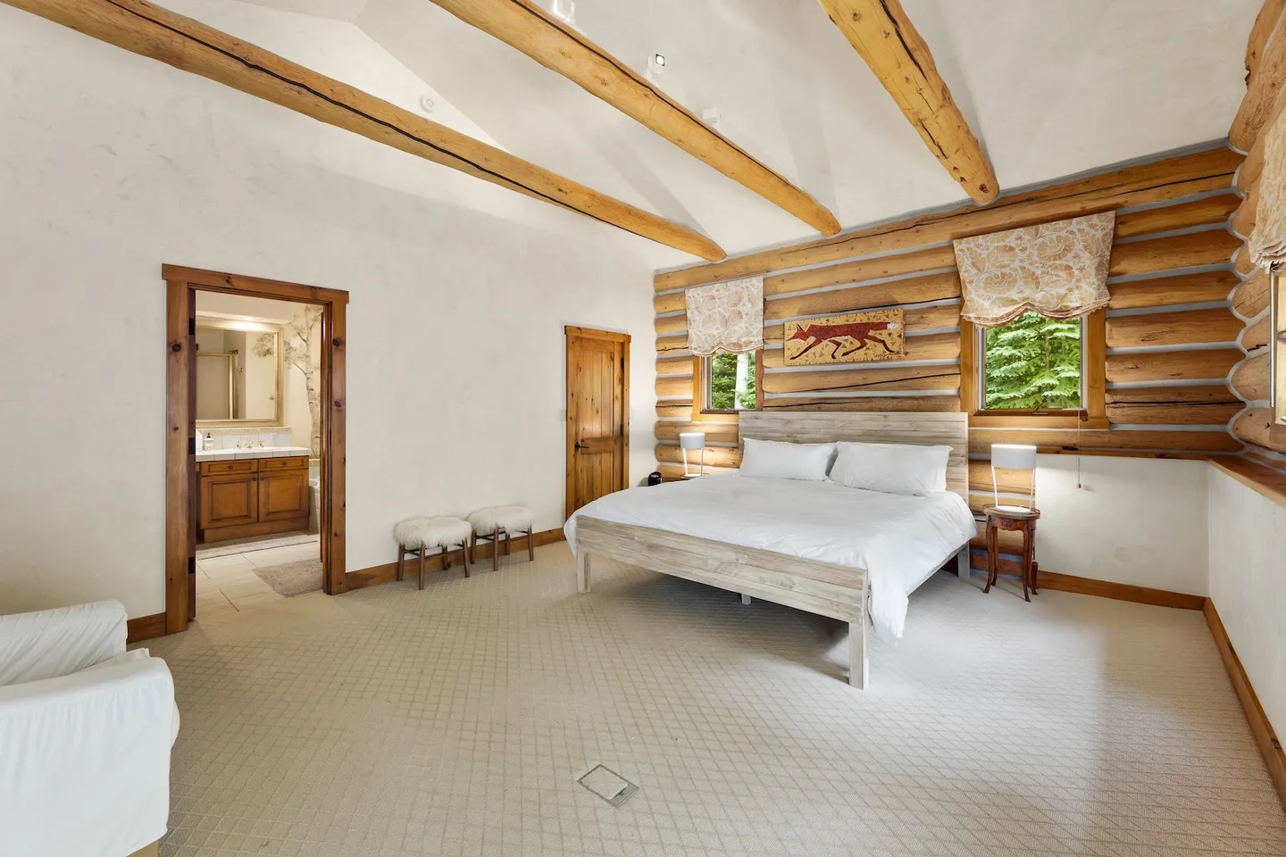 Spacious bedroom with vaulted ceilings, exposed log beams, a log feature wall, and a bed flanked by windows.