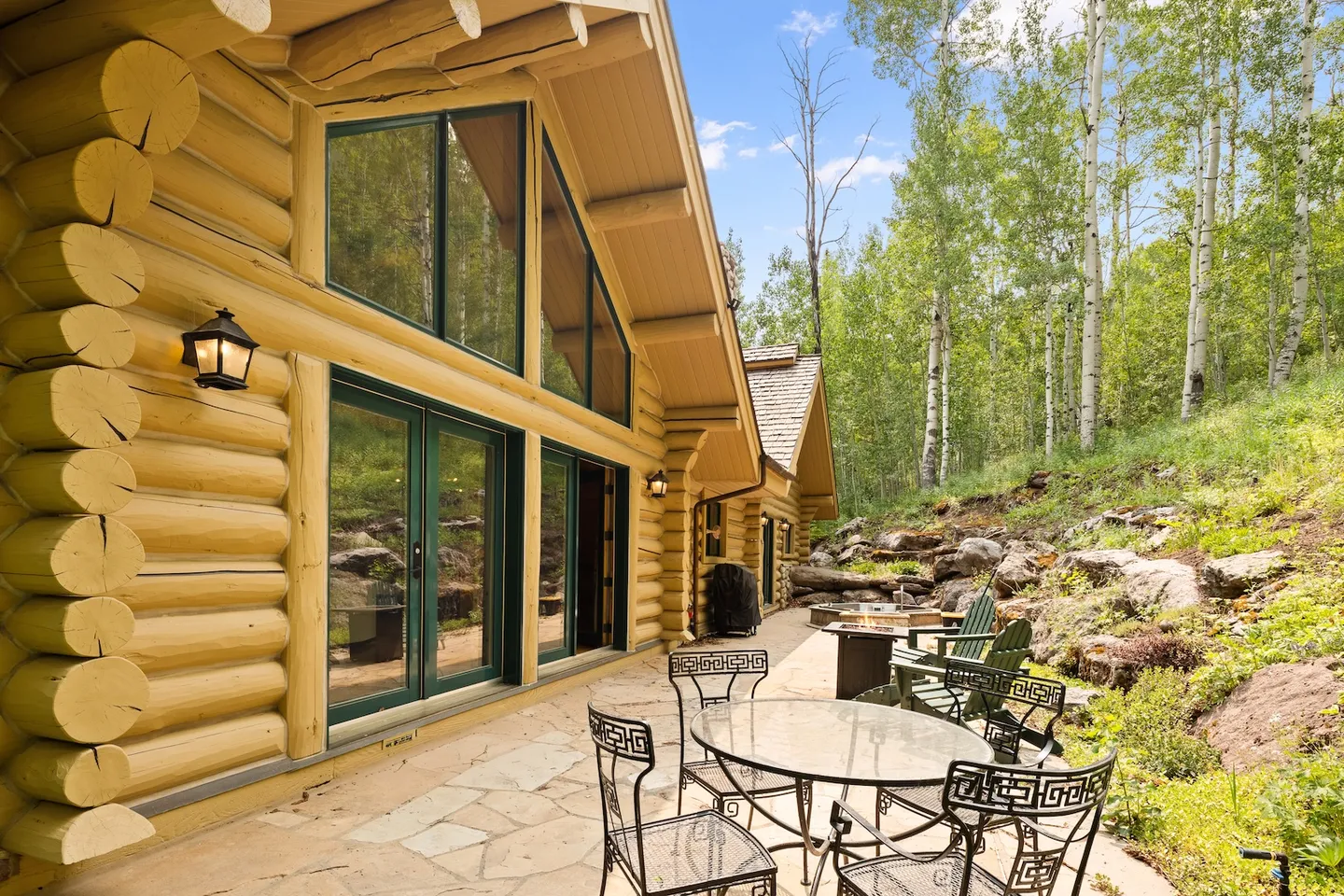 Stone patio alongside a log cabin with outdoor seating, fire pit, and surrounding aspen trees.