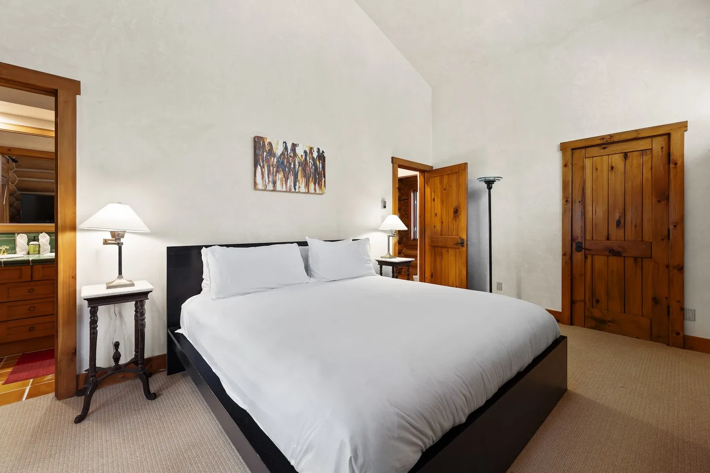 Bedroom with high vaulted ceiling, dark wood bed frame, white bedding, nightstands with lamps, and rustic wooden doors.