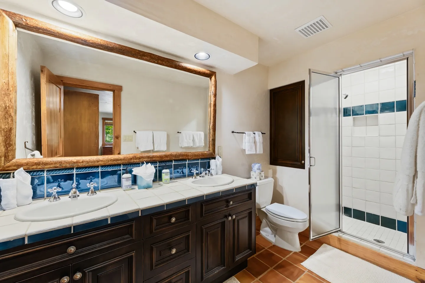 Bathroom with double vanity, tiled countertops, wooden mirror frame, toilet, and walk-in shower with glass door.