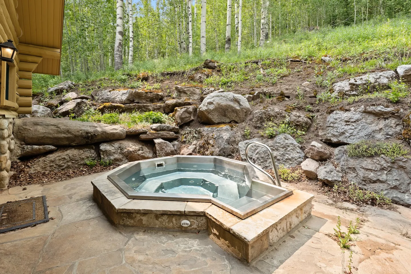 Outdoor hot tub embedded in a stone patio next to a log cabin and a rocky hillside with aspen trees.