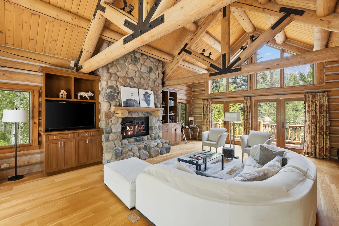 Rustic living room with high vaulted log-truss ceilings, a large stone fireplace, and a curved sectional sofa.
