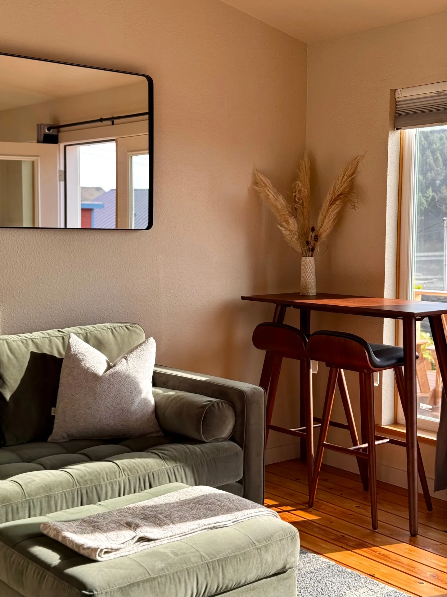 Living room corner featuring a green velvet tufted couch, an ottoman, and a tall wooden bar table with two barstools next to a bright window.