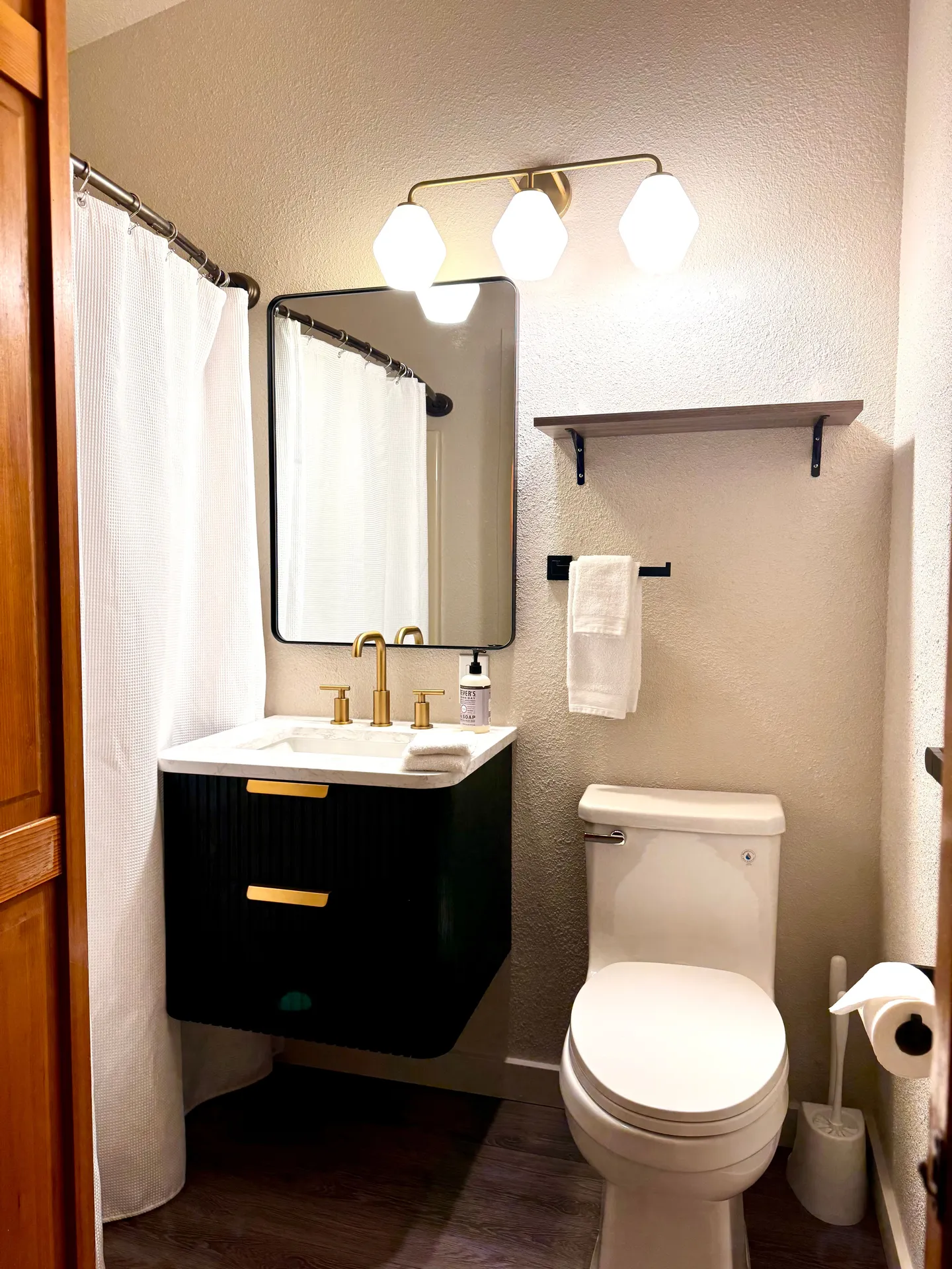 Bathroom featuring a floating black vanity with gold fixtures, a modern mirror, a three-bulb vanity light, and a white toilet.
