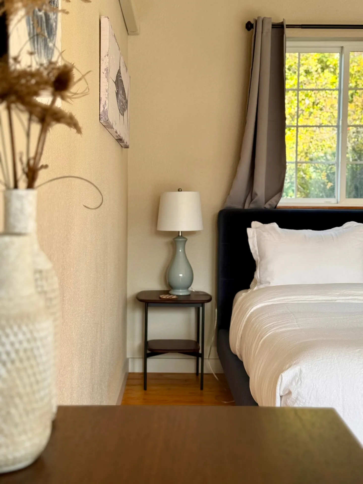 Bedroom detail showing a bed with white linens and an upholstered headboard next to a nightstand with a lamp and a window.