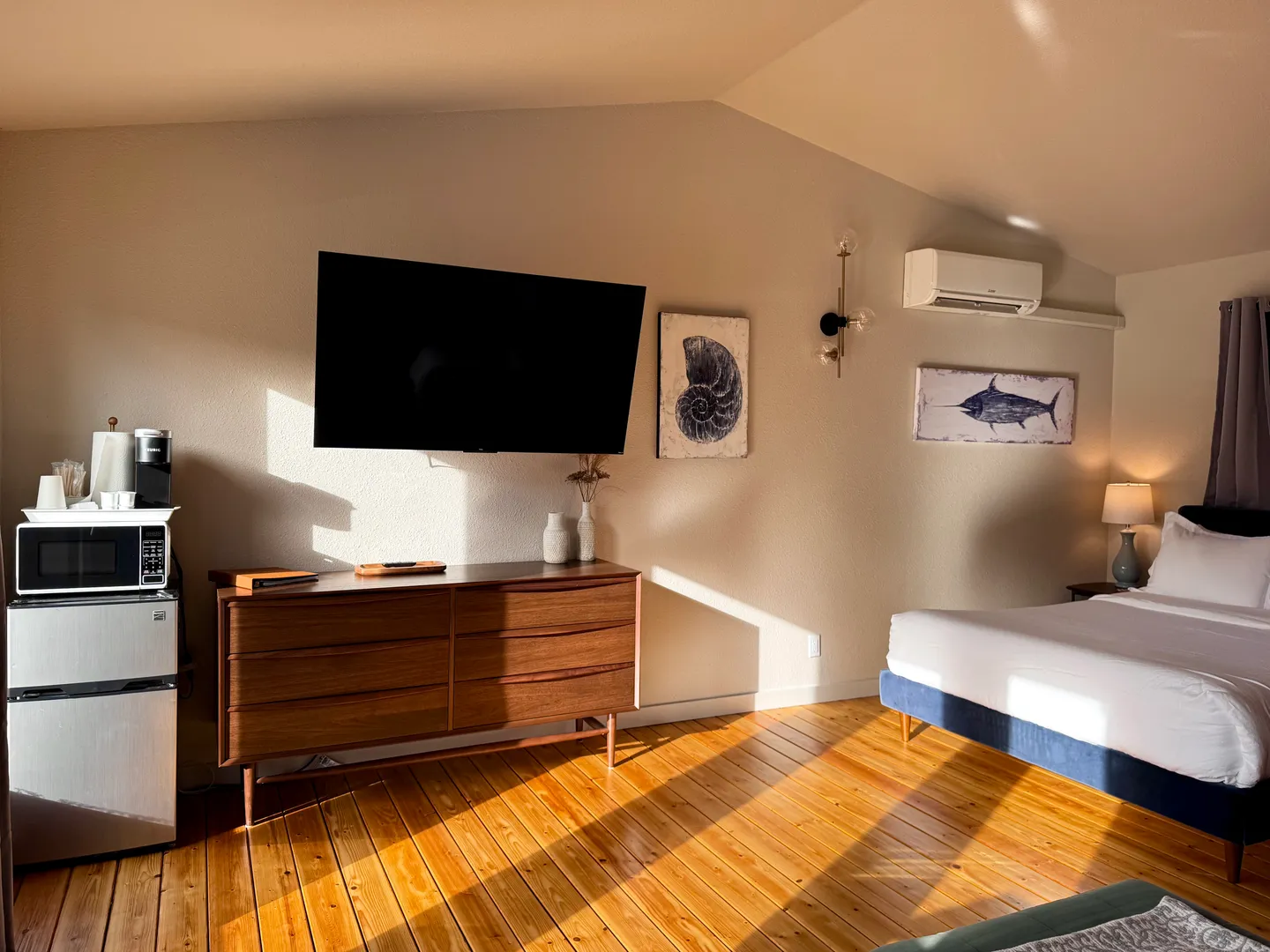 Bedroom featuring wood plank flooring, a wall-mounted TV over a wooden dresser, a mini fridge with microwave, and a bed.