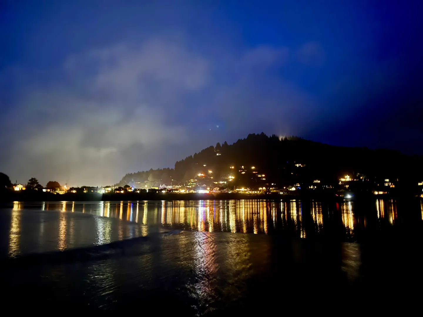 Nighttime view of a coastal hillside dotted with illuminated houses reflecting across a body of water.