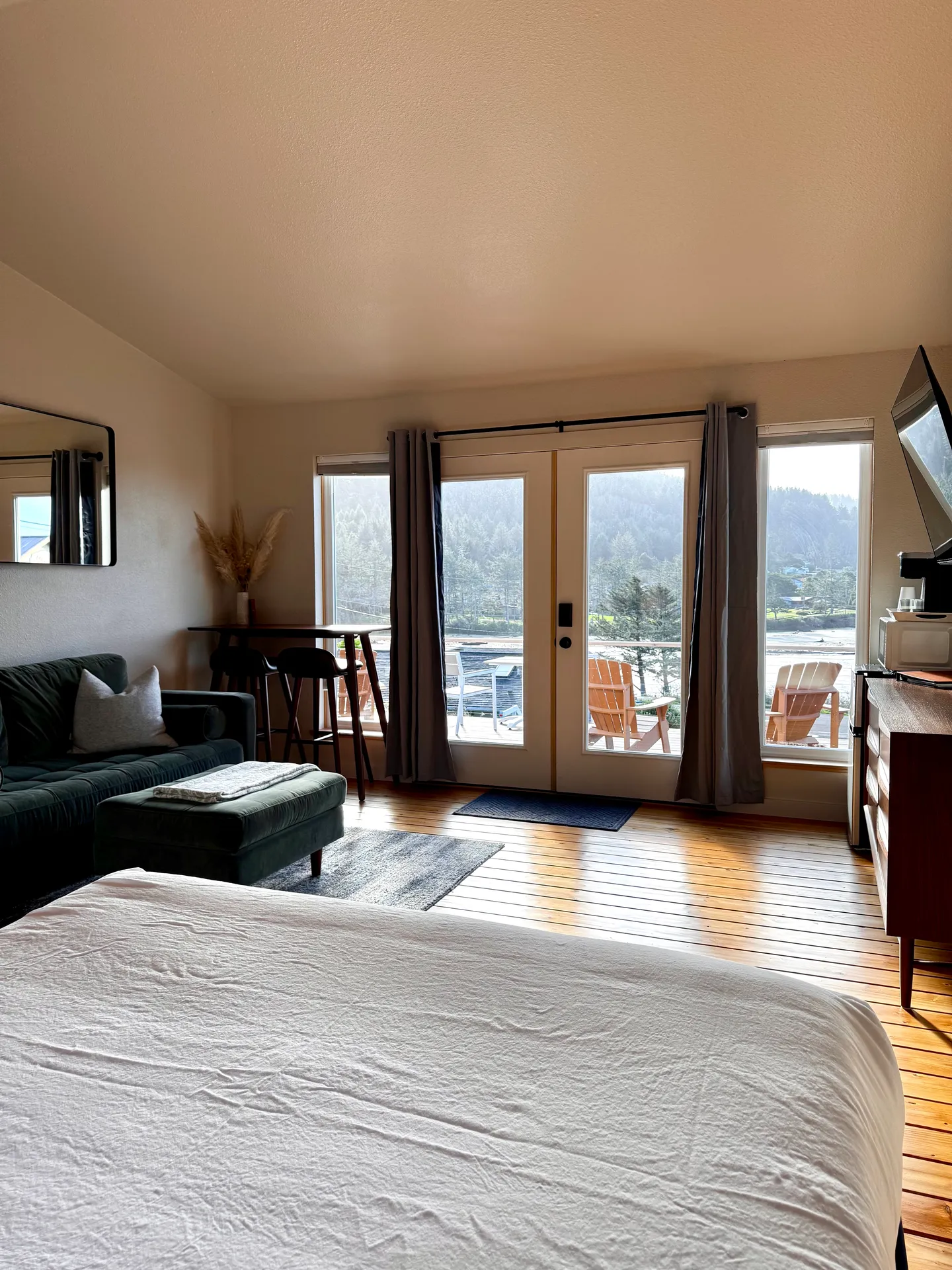 Bedroom with wooden floors, a bed in the foreground, small sitting area, and glass doors opening to a deck overlooking water.