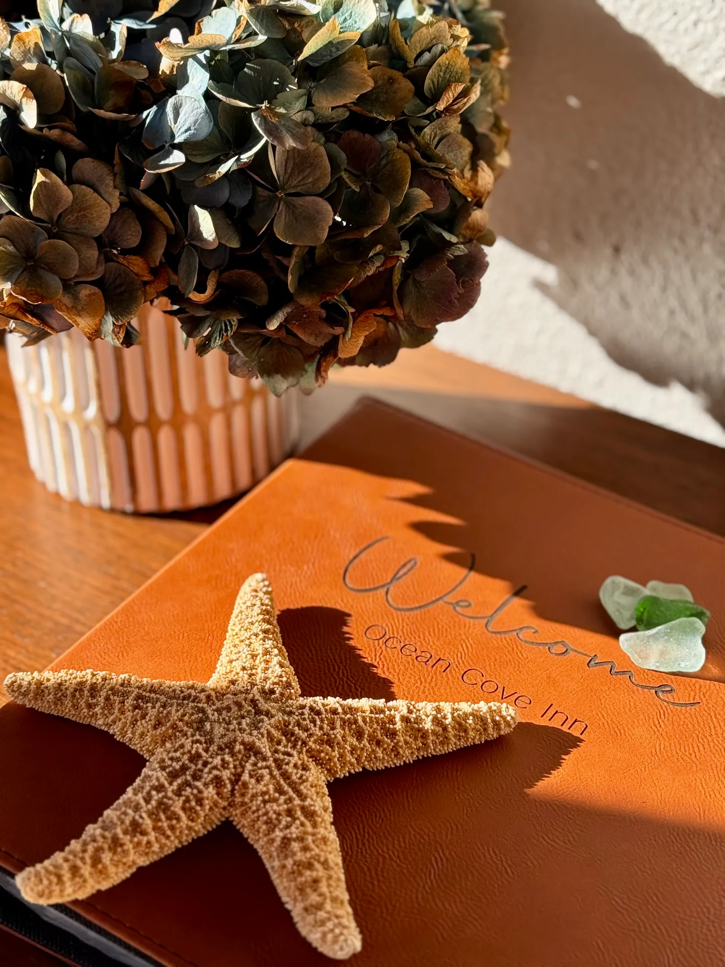 Close-up of a leather welcome binder labeled Ocean Cove Inn, featuring a dried starfish, pieces of sea glass, and a vase of dried flowers.