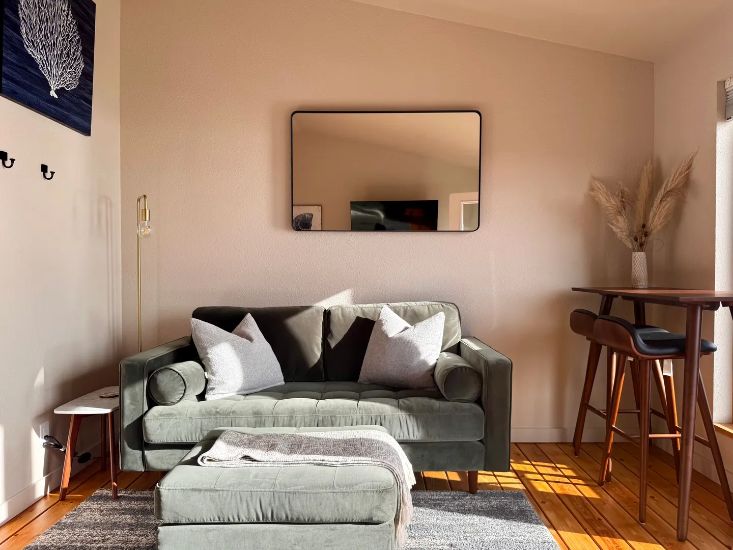 A living area with a green velvet loveseat, matching ottoman, a rectangular wall mirror, and a small wooden high-top table with barstools.