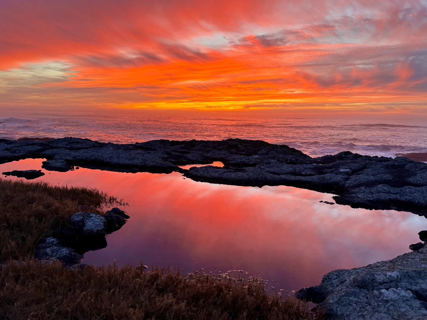 A tide pool in the foreground reflecting a vibrant orange and pink sunset over the ocean horizon.