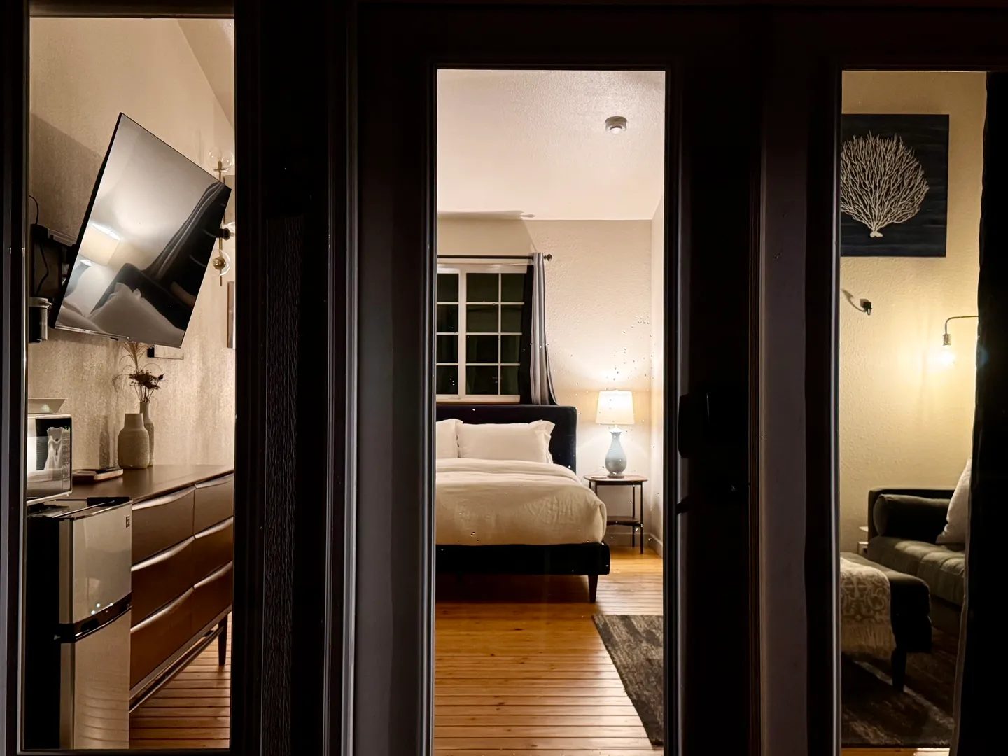 A nighttime view looking through glass-paned doors into an illuminated bedroom with a bed, dresser, TV, and small seating area.