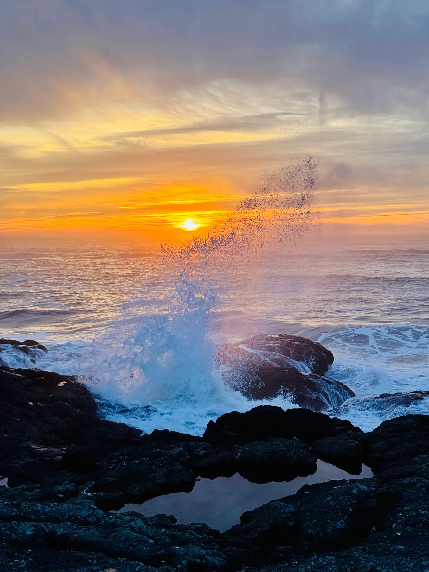 Ocean wave crashing against dark rocks with water spraying upward during a vibrant sunset over the horizon.