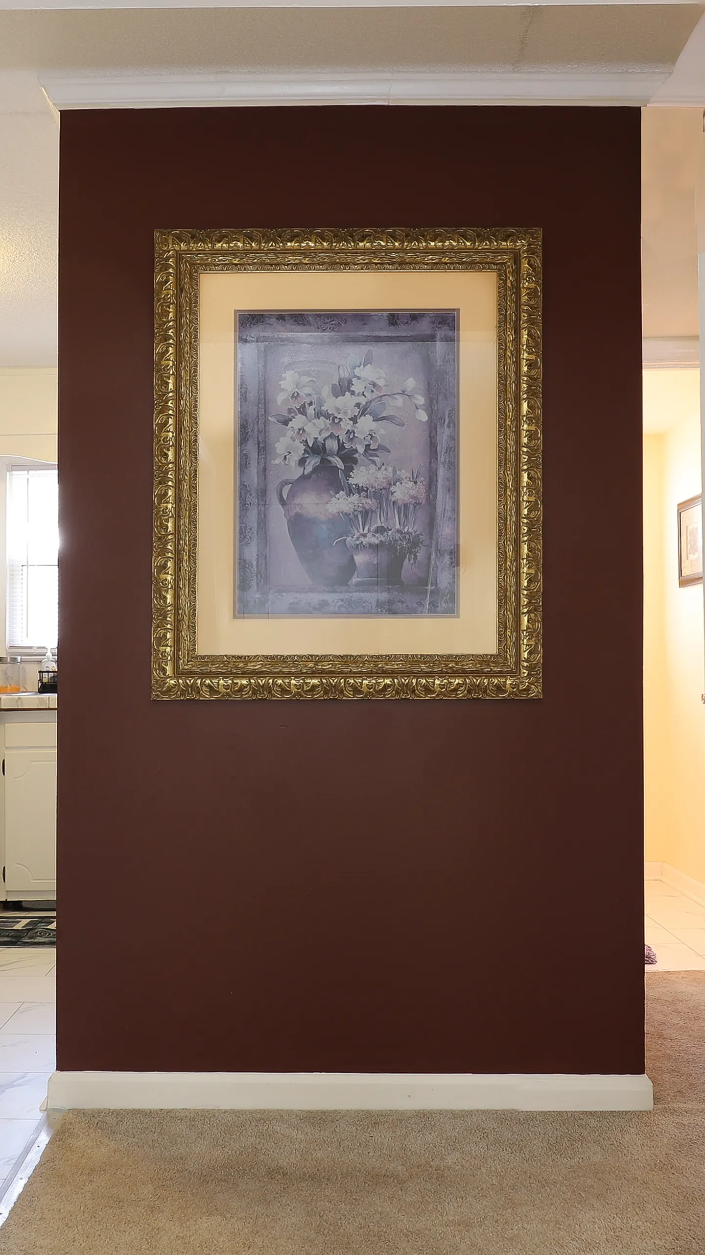 Framed floral artwork in an ornate gold frame hanging on a deep brown accent wall with carpet below.