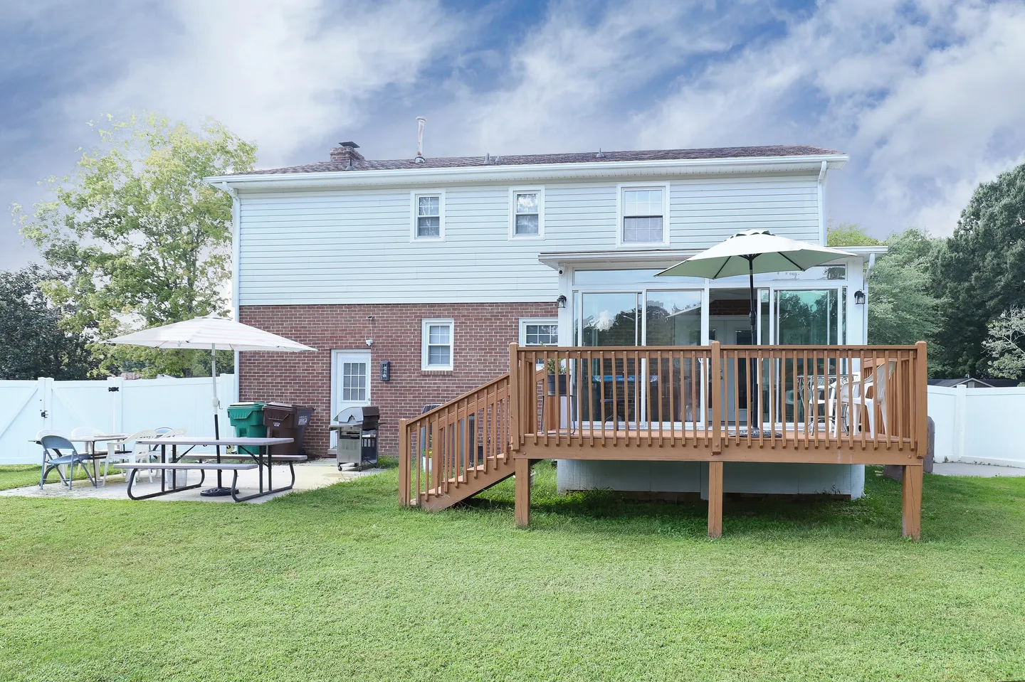 Rear exterior of a two-story home with a green lawn, wooden deck, sunroom, and patio with outdoor dining furniture.