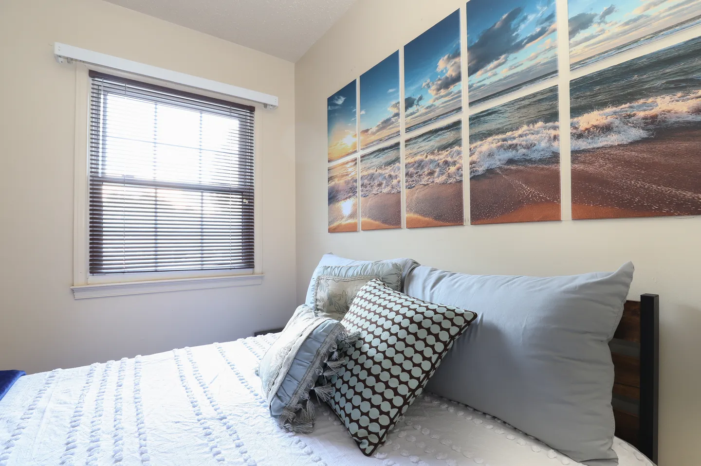 Close-up of a bed with light blue and patterned pillows next to a window with blinds and a multi-panel ocean wave artwork above the headboard.