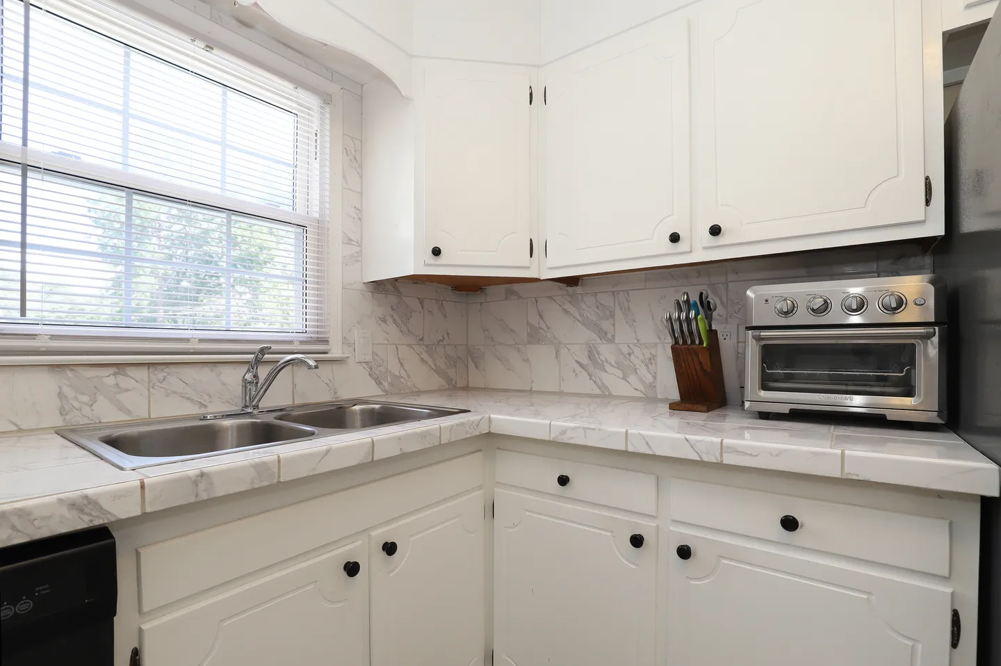 Kitchen corner featuring a double-basin stainless steel sink under a window, white tile countertops with matching backsplash, and white cabinets.