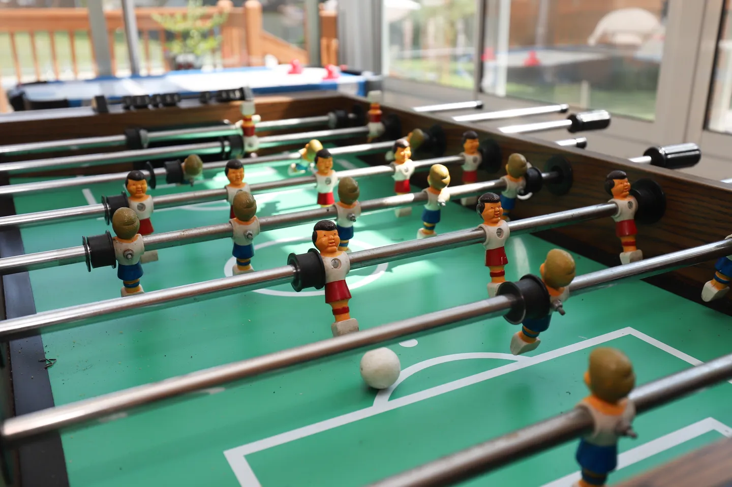 Close-up view of a foosball table with painted player figures and a white ball on a green field indoors near windows.
