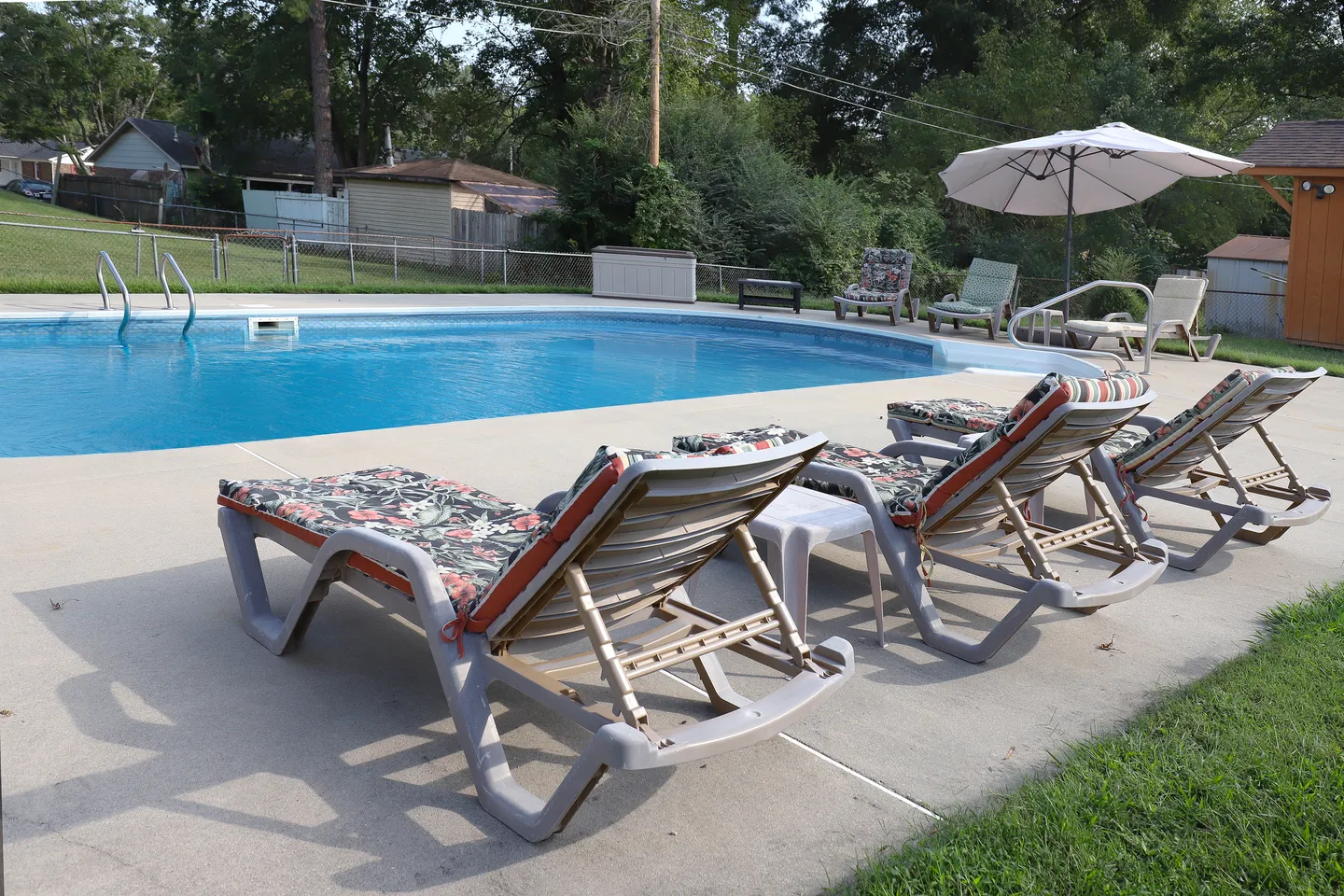 Outdoor swimming pool surrounded by a concrete patio with cushioned lounge chairs and a chain-link fence in the background.