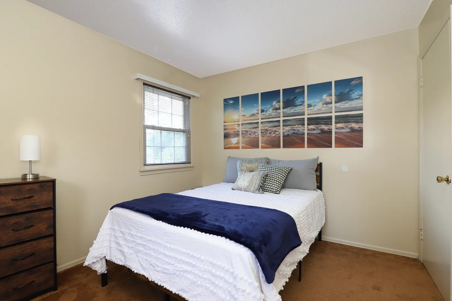 Bedroom with a made bed, wall art depicting a beach sunset, a window with blinds, and a dark wood dresser.