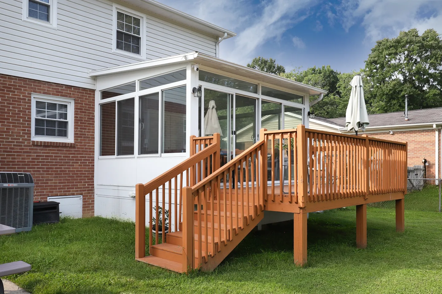 Exterior backyard view showing a wooden elevated deck with stairs connected to an enclosed sunroom with glass windows and sliding doors.