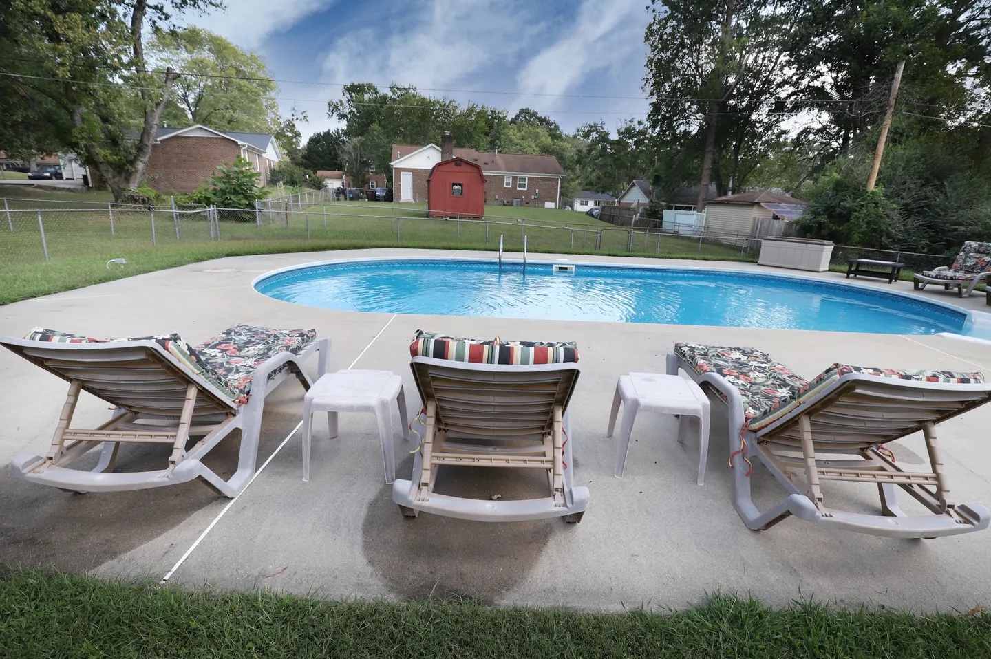 Outdoor swimming pool surrounded by a concrete patio with lounge chairs and a grassy fenced yard in the background.